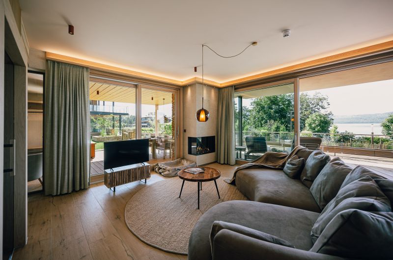 Blick in den Wohnbereich SeeTerrain mit Sofa, Fernseher, Kamin und großen Panoramafenstern mit Blick nach draußen