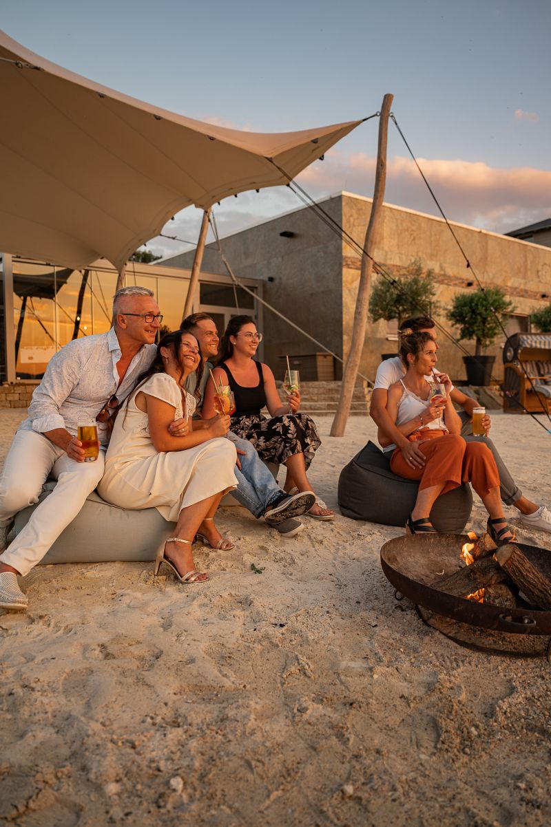 Gruppe sitzt bei Sonnenuntergang am Strand um eine Feuerstelle mit Cocktails in der Hand