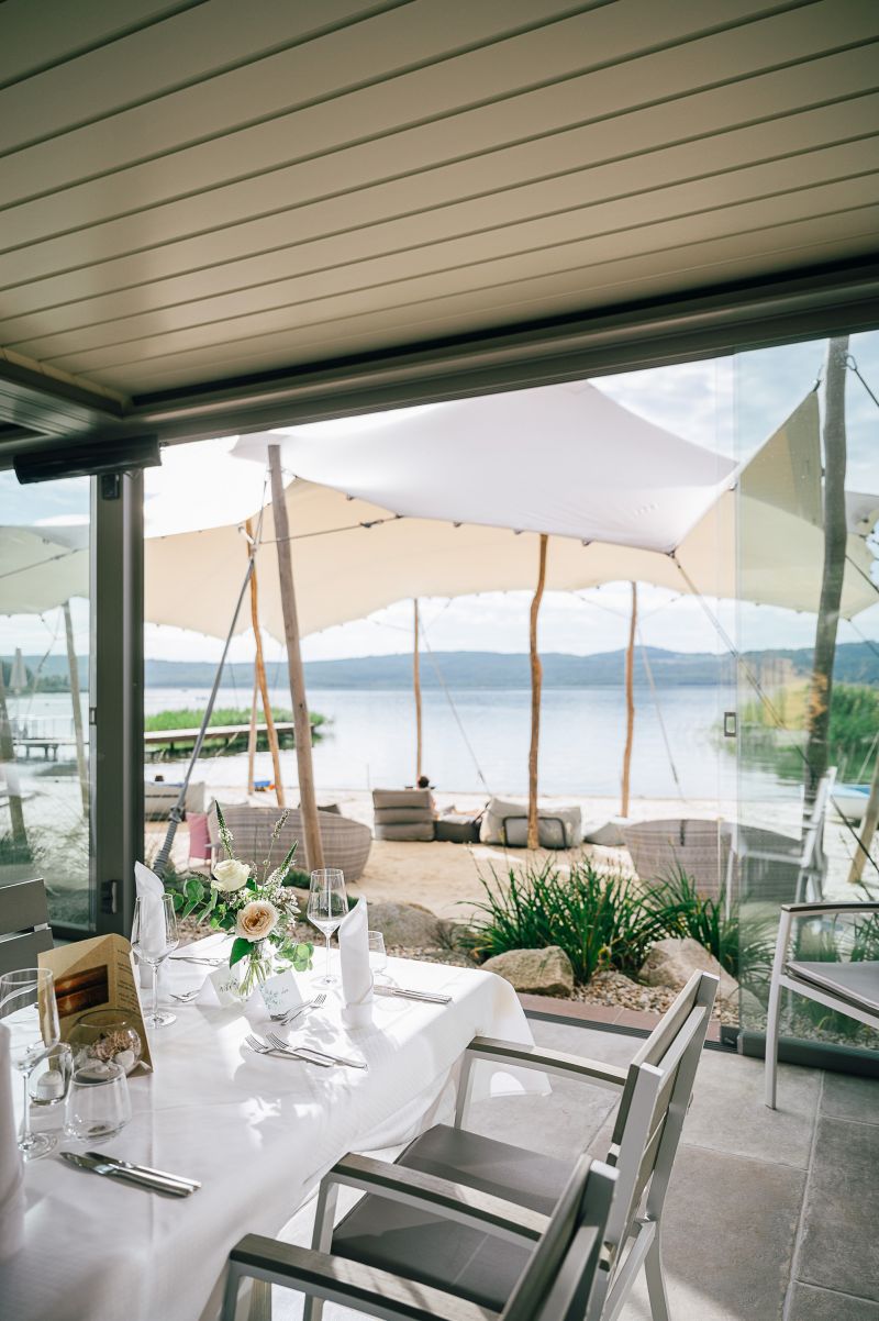 Eingedeckte Tische für eine Feier im Glaspavillon. Im Hintergrund Strand und See